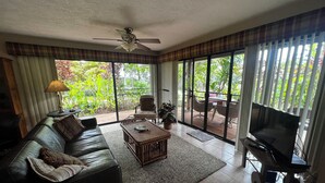 A living room with a sofa, a coffee table, a TV, and a ceiling fan. There is a sliding glass door leading to an outdoor area with a table and chairs. - Molokai condo Pool/Spa, 2 bedrooms/2 bathrooms. Walking distance to the beach.  (Maunaloa)