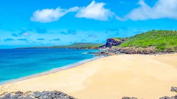 A beach with clear blue water, sandy shore, and a rocky outcrop with greenery. - Molokai condo Pool/Spa, 2 bedrooms/2 bathrooms. Walking distance to the beach.  (Maunaloa)