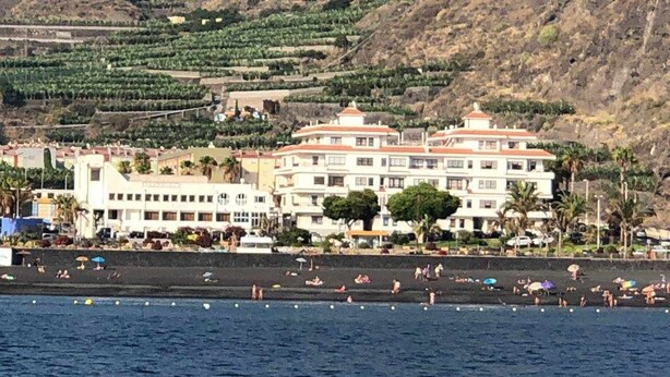 Ein Hotel an der Küste mit mehreren Etagen, ein Strand im Vordergrund und terrassenförmig angelegte Weinberge im Hintergrund.