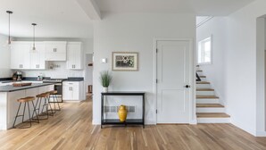 Una cocina moderna con armarios blancos, una encimera negra y taburetes de madera. Hay una mesita con un jarrón y un cuadro en la pared. Una puerta blanca da paso a una escalera.