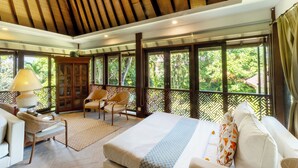 Amplia habitación con una cama grande, un sofá y una silla. Se ve una ventana con vistas a árboles y un techo de madera.