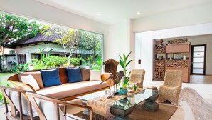 Un salón amplio con un sofá grande, sillas de mimbre, una mesa de centro de vidrio y vistas a una casa tradicional y un jardín exuberante.