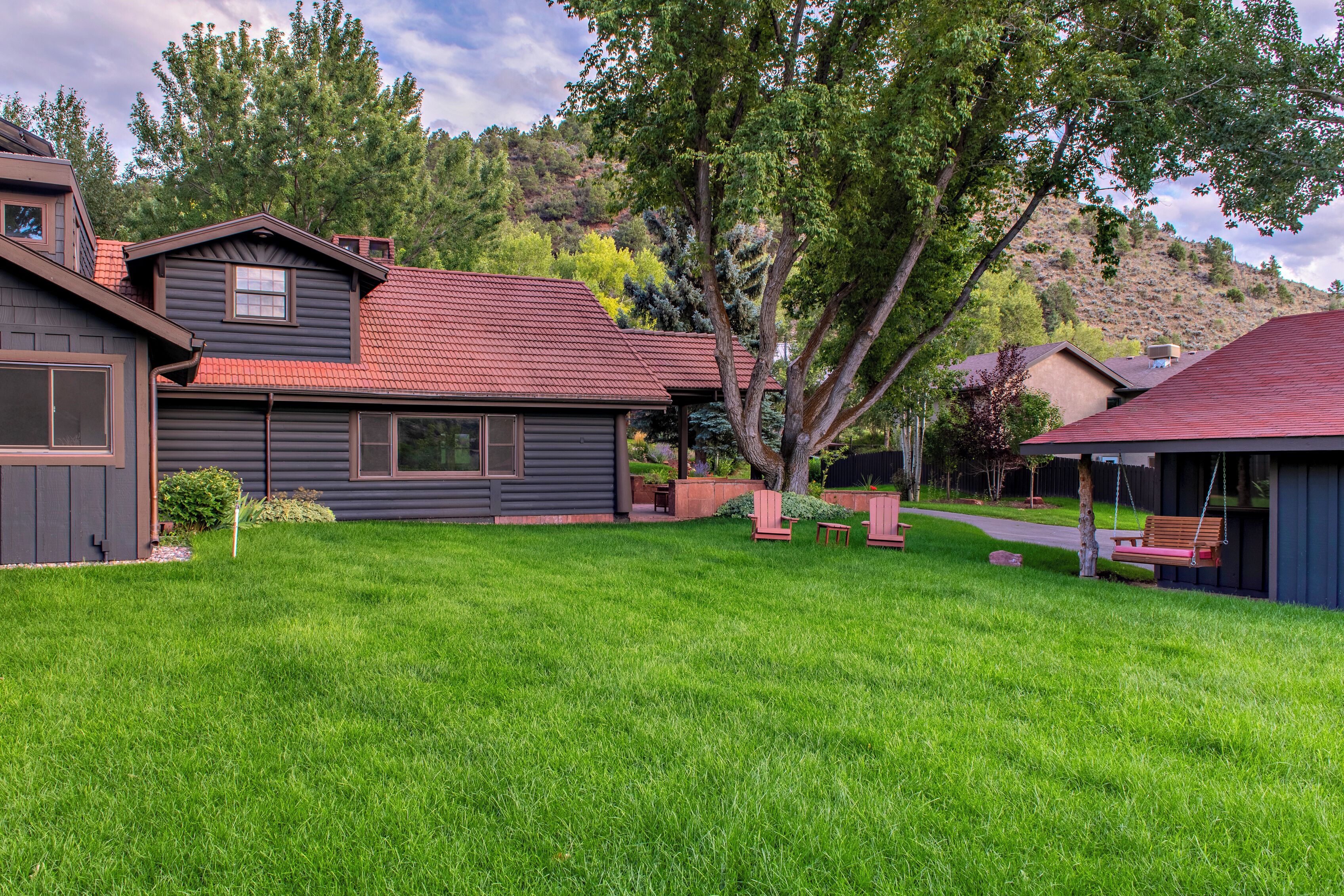 A residential house with a red roof, a front yard with a bench, and a hillside in the background.
