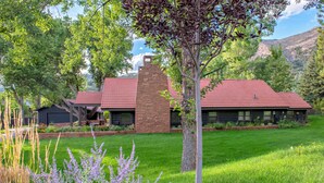 A log cabin with a stone chimney, surrounded by a well-maintained lawn and flowering plants.