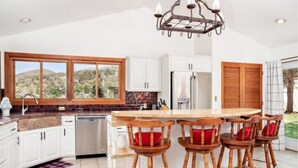 A kitchen with white cabinets, a wooden island with bar stools, a stainless steel refrigerator, and a large window overlooking a hilly landscape.