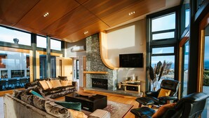 A modern living room with a stone fireplace, leather chairs, and a large window overlooking a scenic view. - Mt Gold Retreat - Tennis court - Hot Tub (Wanaka)