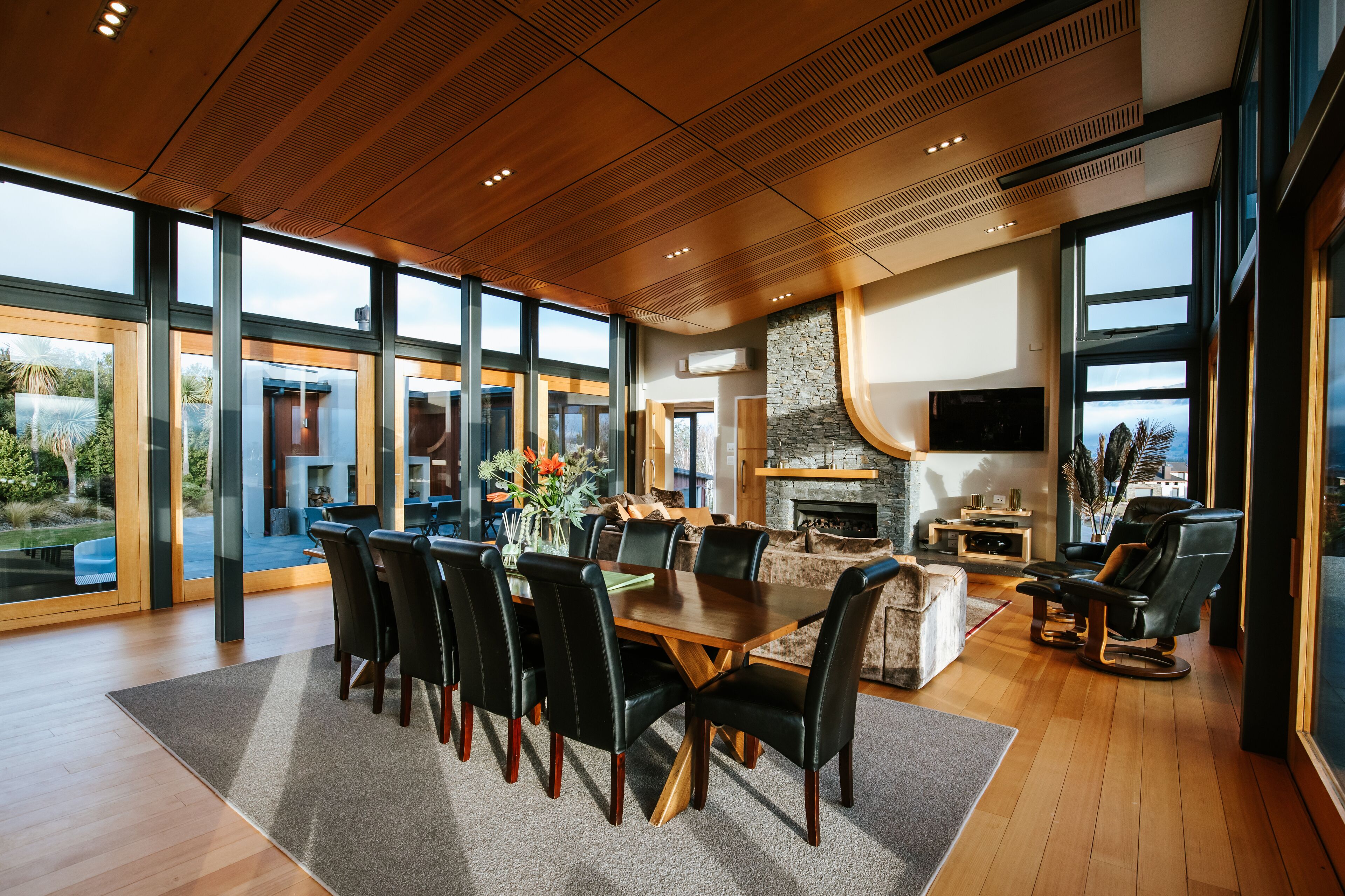 A spacious living area with a wooden dining table, black chairs, a fireplace, and large windows offering a view of the outdoors.