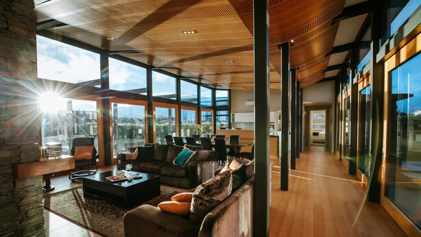A spacious living area with a large window, a fireplace, and a modern kitchen in the background. - Mt Gold Retreat - Tennis court - Hot Tub (Wanaka)
