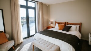 A bedroom with a large bed, a bench, a nightstand, a lamp, and a view through a glass door. - Mt Gold Retreat - Tennis court - Hot Tub (Wanaka)