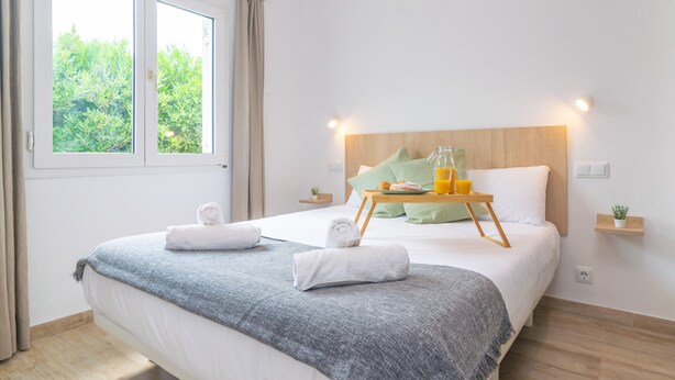 Ein ordentlich bezogenes Bett mit grauer Decke, zwei aufgerollte Handtücher und einem Tablett mit Orangensaft und einem Gebäck.