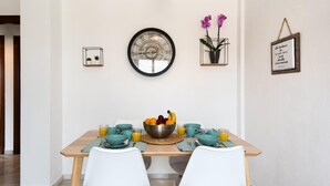 A dining area with a wooden table, two white chairs, a fruit bowl, and a wall clock.