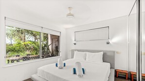 A modern hotel room with a large bed, a window with a view of greenery, a ceiling fan, and a small wooden bench. - 5 mins walk to Hastings Street (Noosa)