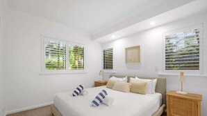 Un dormitorio con cama, dos mesitas de noche, un aparador, una ventana con persianas y una obra de arte colgada en la pared.