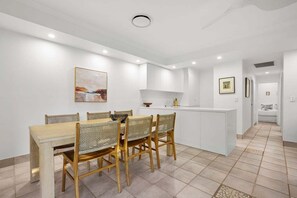 A modern kitchen with white cabinetry, a dining area with a wooden table and wicker chairs, and a hallway leading to a bedroom. - Noosa River at your doorstep (Noosa)