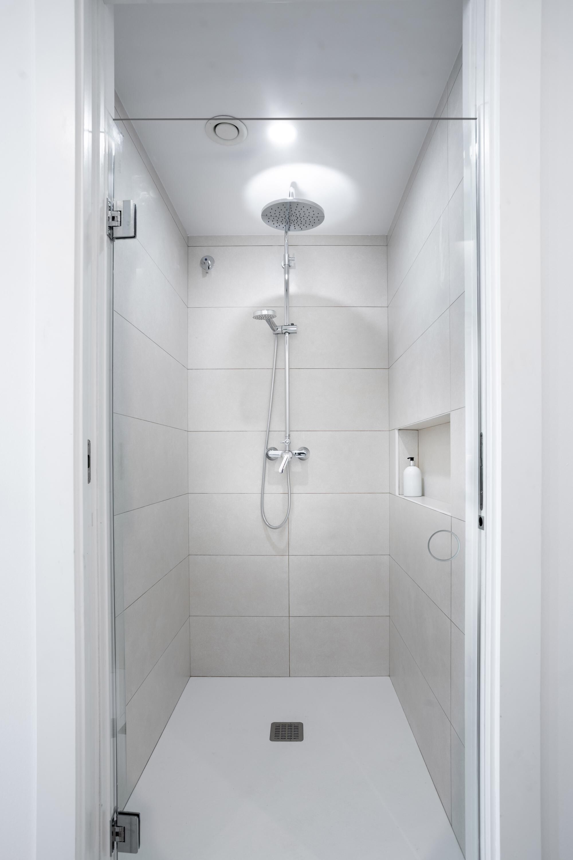 Eine moderne Dusche mit Handbrause und Deckenbrause, einem kleinen Ablagefach mit einer Flasche sowie einem Fußbodenlüfter.