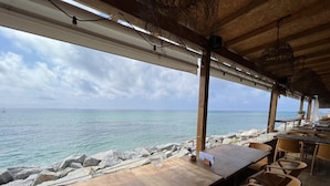 Ein Essbereich im Freien mit Holztischen und -stühlen mit Blick auf ein ruhiges Meer und eine felsige Küste.