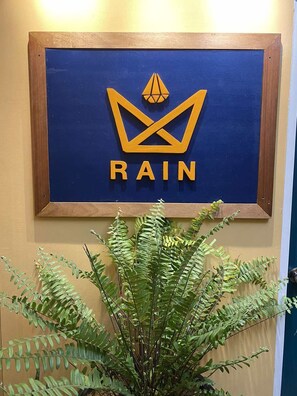 Un cartel enmarcado con la palabra 'RAIN' y un símbolo de corona, colgado en una pared con una planta al frente.