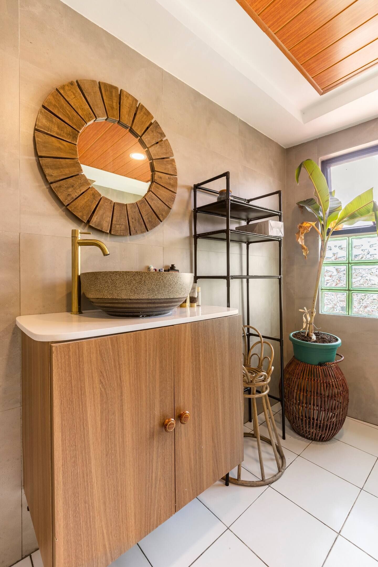 A bathroom with a wooden vanity, a round mirror, a sink with a gold faucet, a potted plant, and a window with a view.
