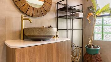 A bathroom with a wooden vanity, a round mirror, a sink with a gold faucet, a potted plant, and a window with a view.