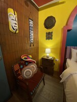 Una habitación con pared de madera, pared amarilla, cama con ropa blanca, mesita de noche con lámpara y silla con cojín colorido.