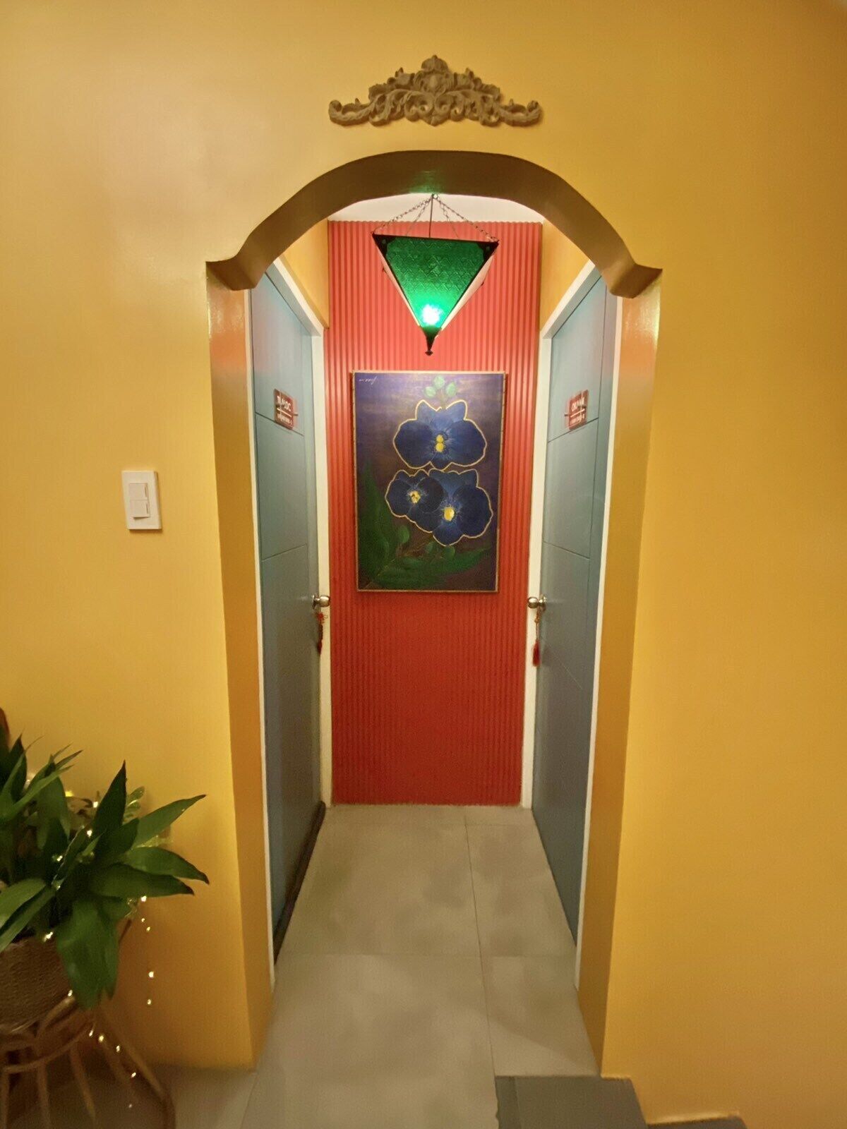 Un pasillo con una puerta roja, una luz verde arriba y un cuadro floral en la pared.