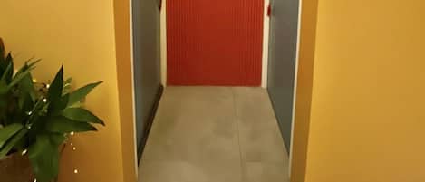 Un pasillo con una puerta roja, una luz verde arriba y un cuadro floral en la pared.