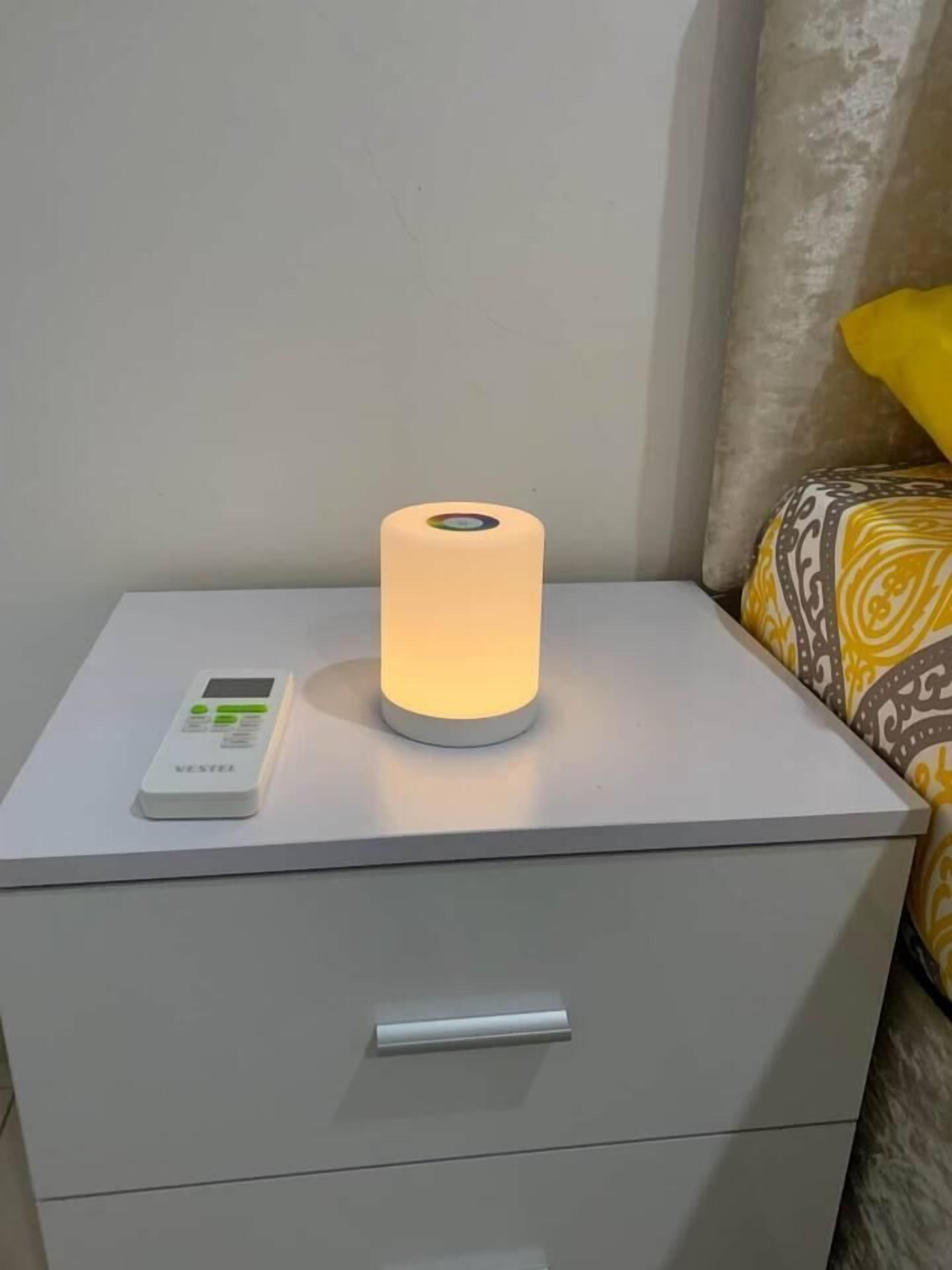 Een nachtkastje met een witte nachtlampje, een cilindervormige lamp en een digitaal apparaat erop.