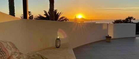 Un espacio al aire libre con asientos curvos, un sofá y cojines, un farol y vista al atardecer sobre el océano.