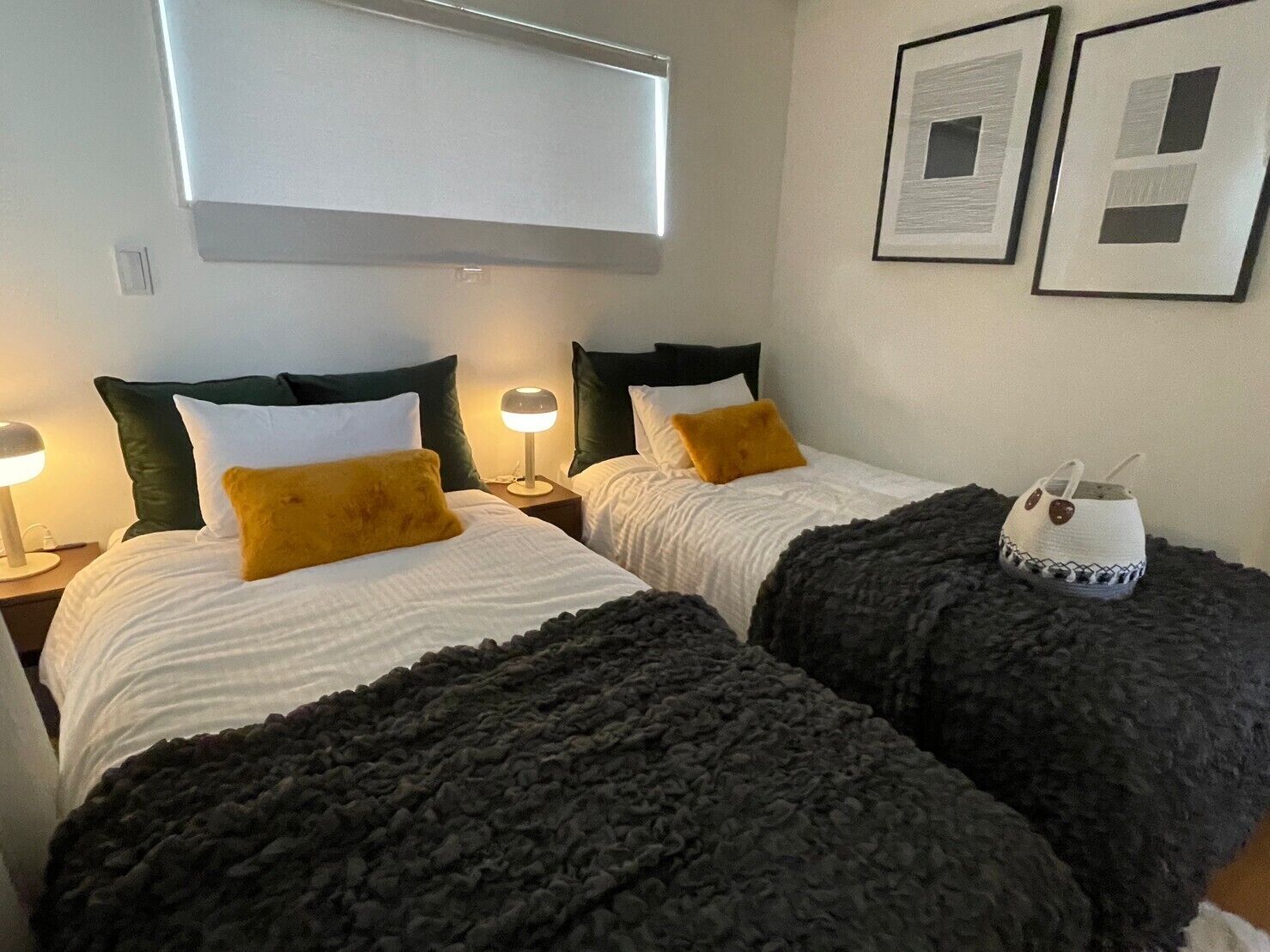 A hotel room with two beds, each with a white comforter and dark throw blankets. There are two bedside lamps, a window with blinds, and two framed pictures on the wall.