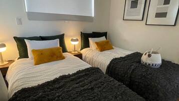 A hotel room with two beds, each with a white comforter and dark throw blankets. There are two bedside lamps, a window with blinds, and two framed pictures on the wall.