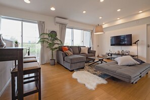 Uma sala de estar moderna com sofá, poltrona e mesa de centro, uma TV na parede e uma varanda com planta.