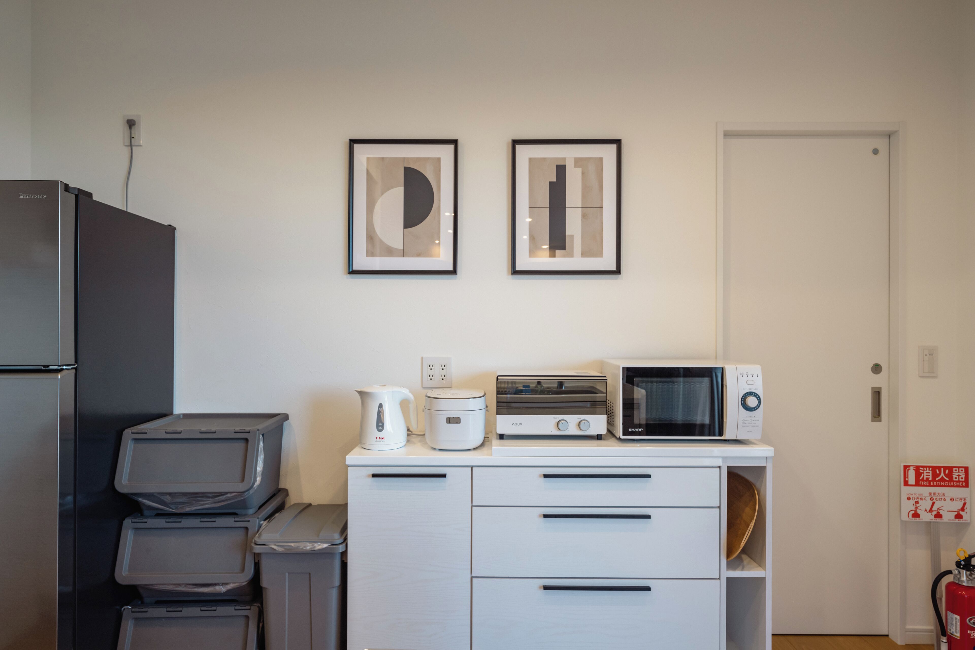 A compact kitchen with a refrigerator, microwave, toaster, and kettle on a white countertop. There are two framed abstract artworks on the wall.