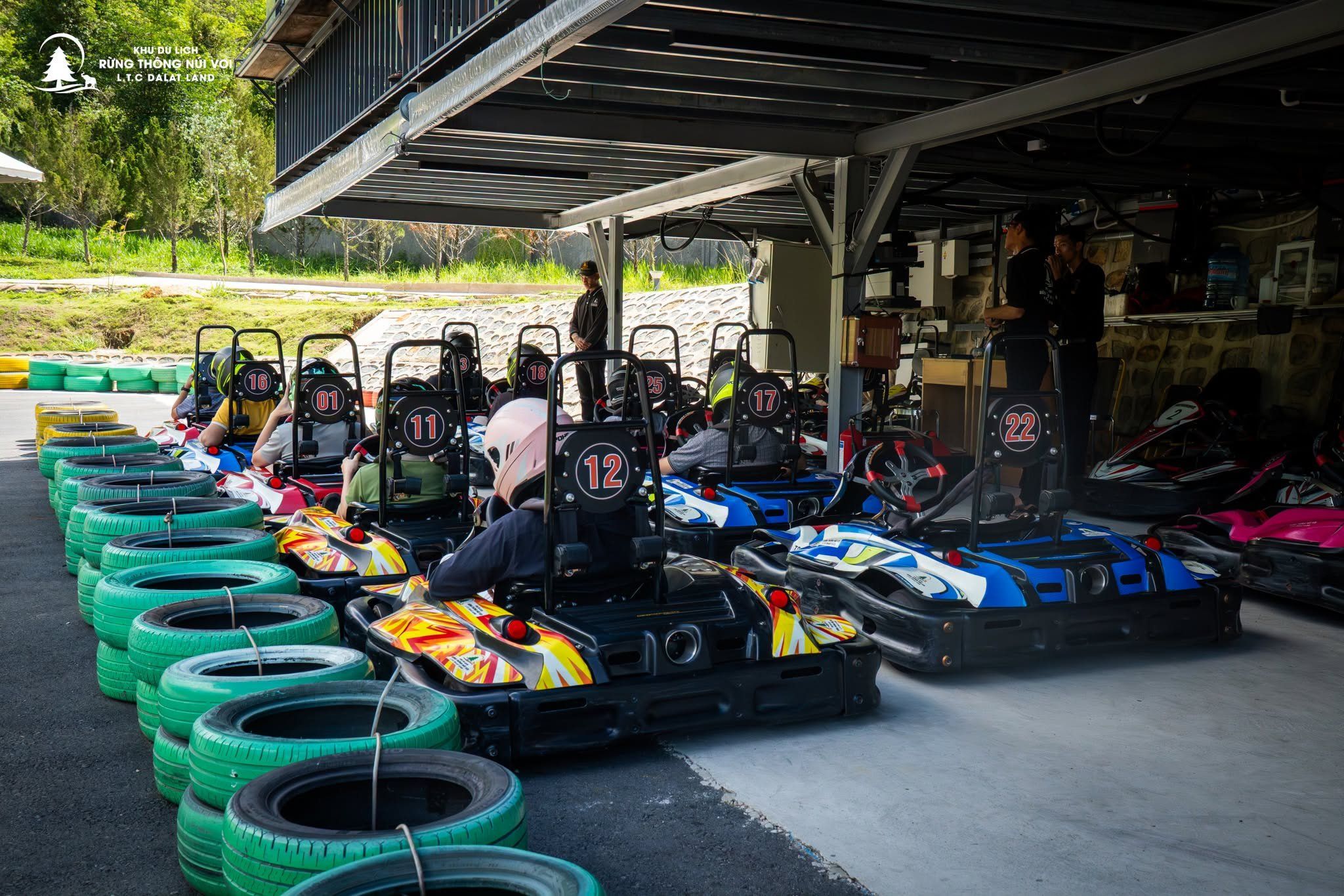 Go-karts lined up with drivers ready to race, a covered area with a roof, and a building with a sign.