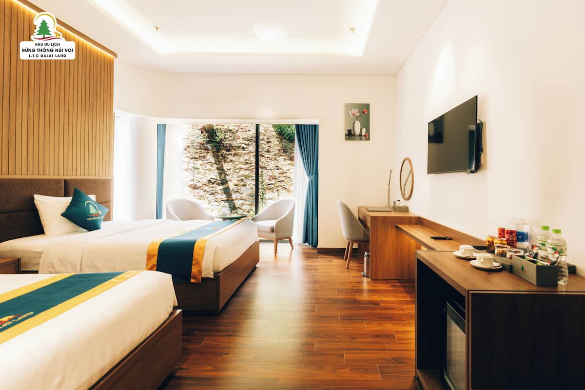 Ein Hotelzimmer mit zwei Betten, einem Schreibtisch, einem Stuhl, einem Fernseher und einem Fenster mit Blick auf Bäume.