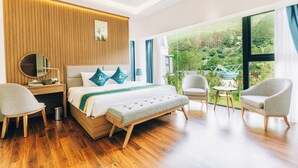 Ein modernes Hotelzimmer mit einer Wandpaneinung aus Holz, einem großen Bett mit Kopfteil, zwei Sesseln, einem kleinen Tisch und Blick auf einen üppigen Garten durch große Fenster.