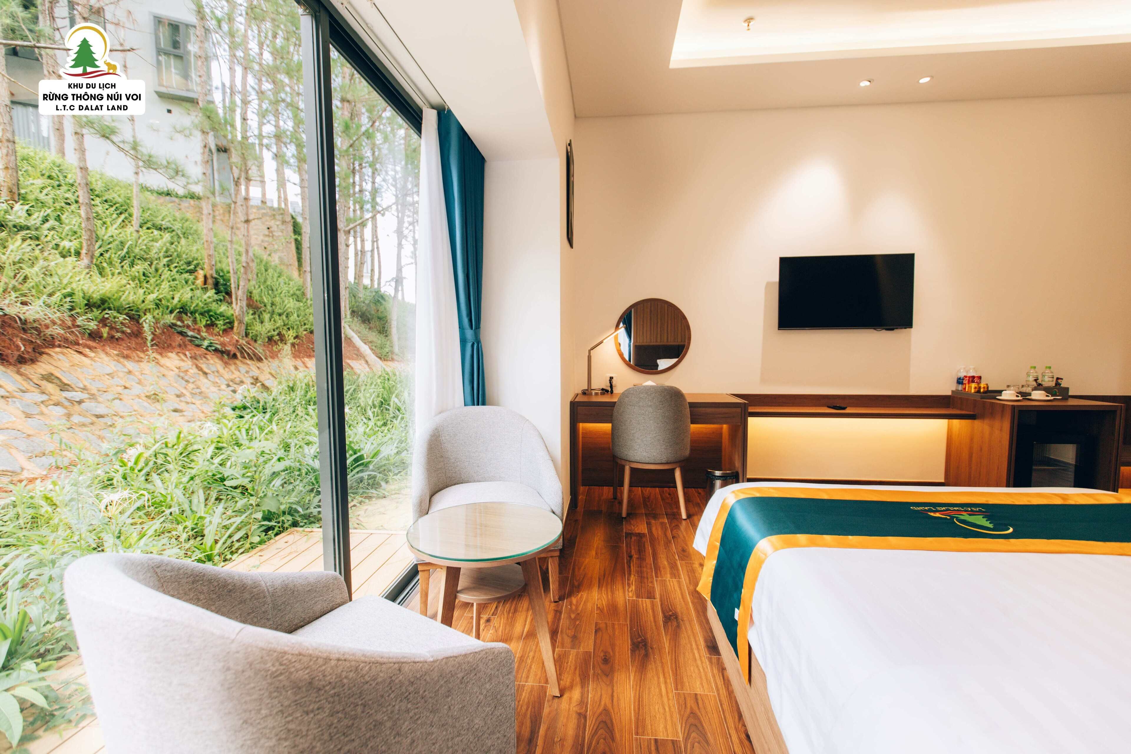 Quarto de hotel com cama, escrivaninha, cadeira, televisão e vista para o verde lá fora.
