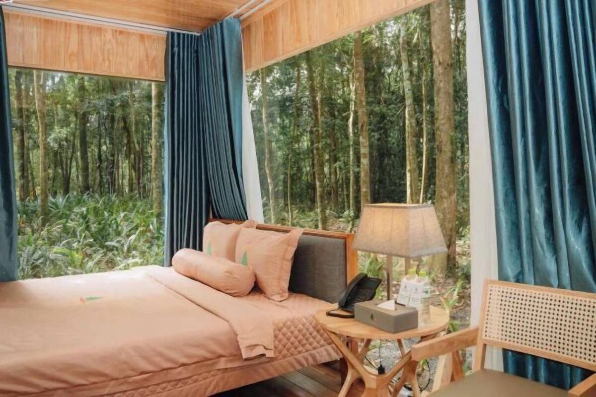 Ein Schlafzimmer mit Bett, Nachttisch, Lampe und Blick auf einen Wald.