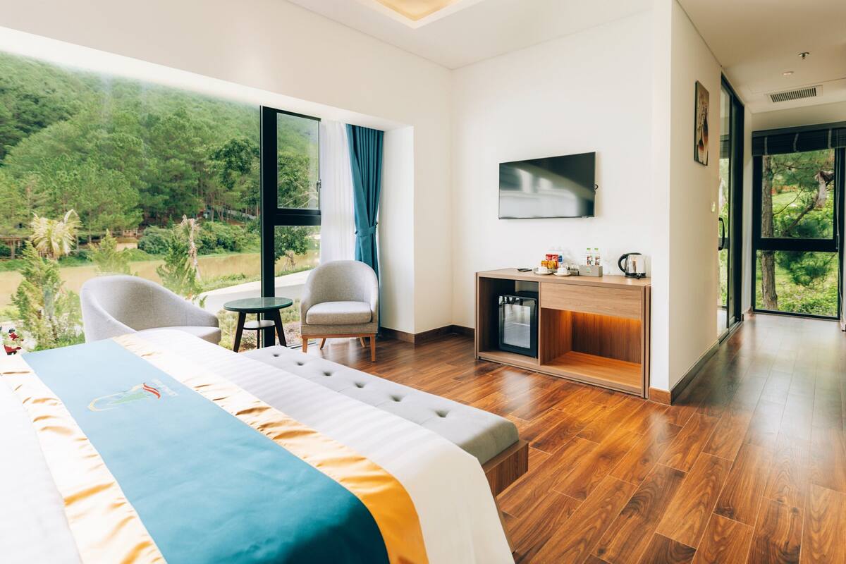 Ein modernes Hotelzimmer mit einem großen Bett, einer Sitzecke mit Stühlen, einem kleinen Tisch, einem Fernseher und Blick auf ein bewaldetes Gebiet.