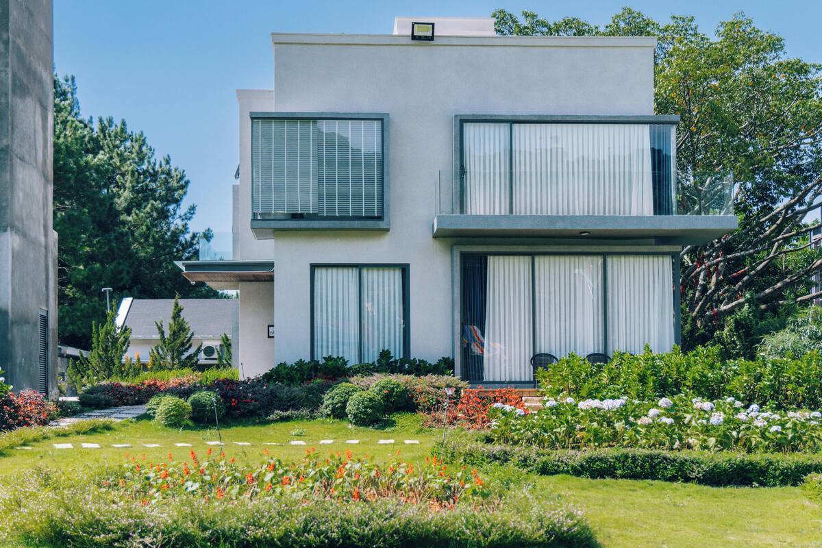 Ein modernes, zweistöckiges Gebäude mit großen Fenstern, ein gepflegter Garten mit verschiedenen Pflanzen und Blumen sowie ein klarer Himmel.