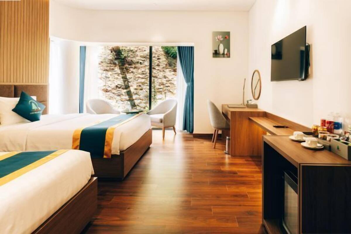 Ein Hotelzimmer mit zwei Betten, einem Schreibtisch, einem Fernseher und Blick auf eine Steinmauer und Grünflächen.