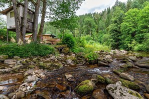 A rustic cabin with a wooden deck and a stone structure, surrounded by dense forest and a flowing stream.