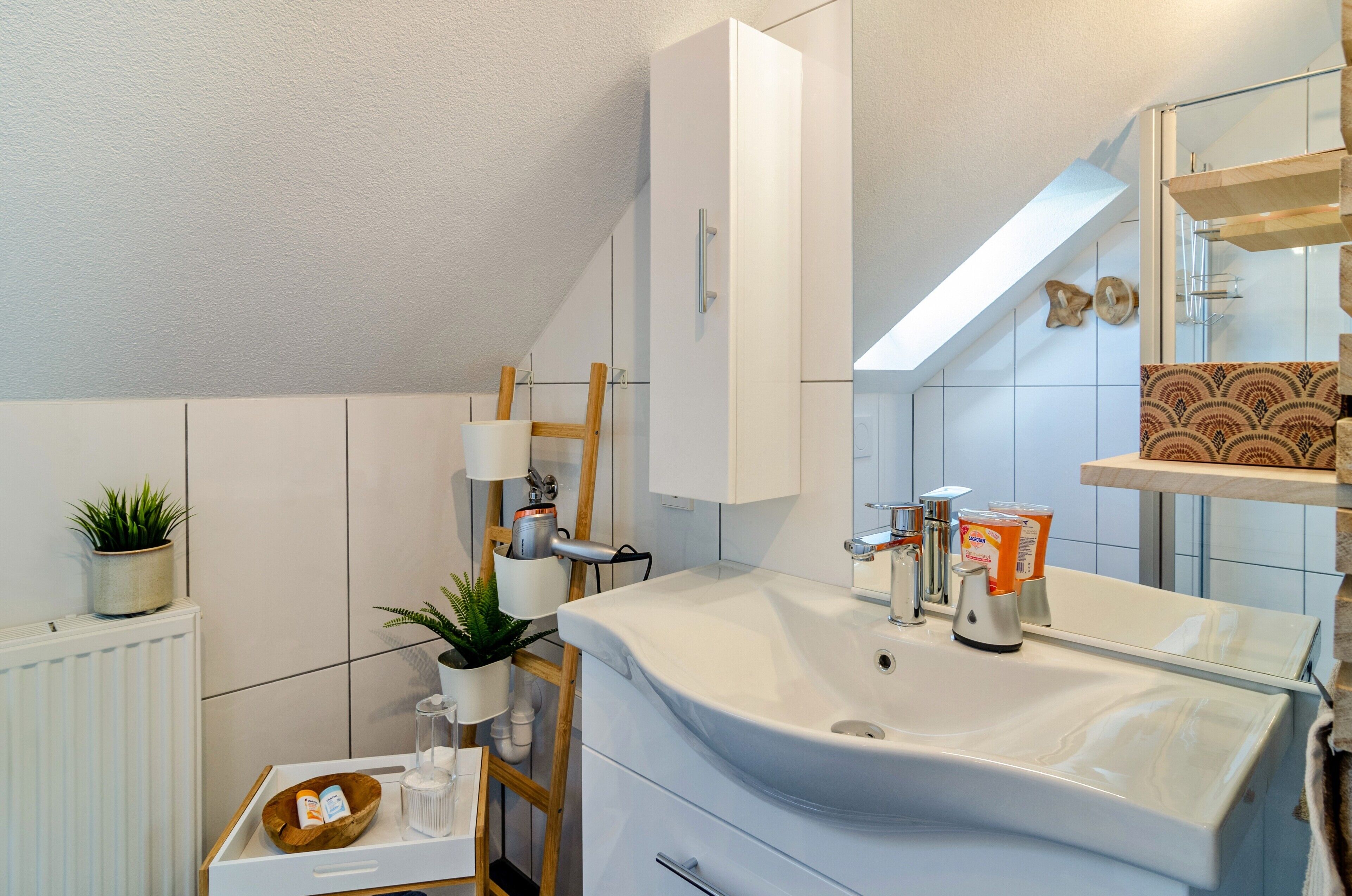 Un baño moderno con un lavamanos, espejo y muebles blancos, un radiador y una planta pequeña.