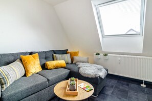 Ein modernes Wohnzimmer mit einem grauen Sofa, gelben Kissen, einem runden Holztisch und Blick auf ein Gebäude durch ein Oberlicht.