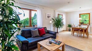 Una sala de estar moderna con sofá, mesa de centro, mesa de comedor y sillas. Hay plantas en macetas y un cuadro en las paredes.