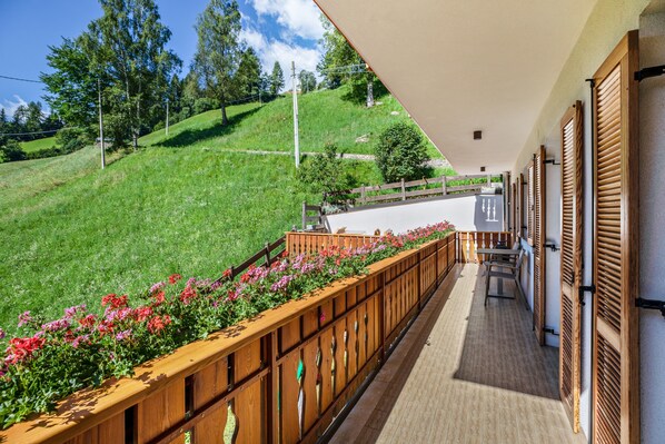 A balcony with a wooden railing, a view of a green hillside, and a table with chairs.