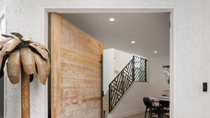 A hallway with a wooden door, a staircase with a geometric railing, and a dining area with a table and chairs. - Urban Beach House in Downtown Huntington Beach (Huntington Beach)