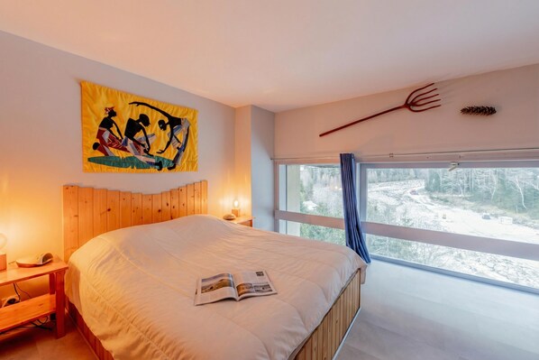 Un dormitorio con cabecera de madera, cama con cubrecama blanco, una revista sobre la cama, mesita de noche con lámpara, ventana con vista a un paisaje nevado y decoración en la pared.