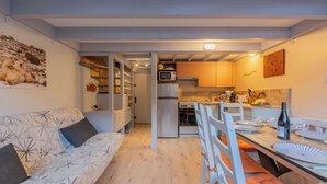 Et kompakt køkken med moderne hårde hvidevarer, et spiseområde med et bord til to, og en hyggelig siddekrog med en sofa.