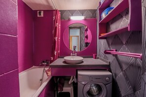 Uma casa de banho compacta com banheira, lavatório, máquina de lavar roupa e espelho com moldura circular.