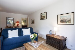 Een woonkamer met een blauwe bank, een houten salontafel en een vaas met bloemen. Er hangen ingelijste foto's aan de muur en staat een lamp op een bijzettafeltje.